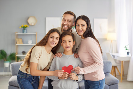 Happy loving smiling family, mother and father with two little kids holding red paper heart, looking at camera, smiling parents and children together, feeling care, emotionally and physically safeの写真素材