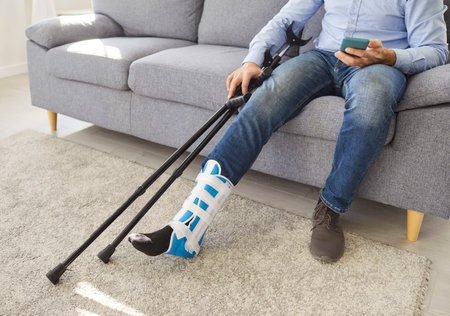 Cropped shot of man with injured leg wearing orthopedic boot sitting on sofa in living room with crutches resting beside him as he using smartphone, recovering and relaxing at home after foot trauma.の写真素材
