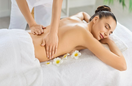 Pleased woman lying with closed eyes on massage table while professional masseuse is providing back massage at luxurious spa, feeling relaxed and comfortable, enjoying peacefulnessの写真素材