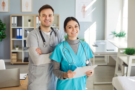 Happy medical staff at work in clinic or hospital. Portrait of 2 smiling doctors, young man and woman general practitioner and intern in scrubs with clipboard, standing in office and looking at cameraの写真素材