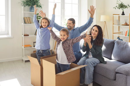 Happy family celebrating purchase of new home and relocation with kids standing in cardboard boxes in living room. Cheerful parents and children raising hands in excitement during moving day fun.の写真素材