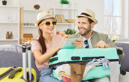 Happy cheerful couple in beach hats sitting on sofa at home with packed suitcase making a shopping list for vacation. Man and woman going on holiday trip. Summer journey and travelling concept.の写真素材
