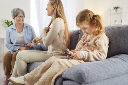 Little child girl using mobile phone while her grandmother talking with mother sitting on sofa in living room at home. Kid with smartphone resting on couch with family. People and technology.の写真素材