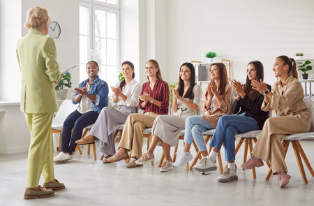 Confident mature woman giving presentation to group of young attentive women sitting in row in bright room. Multiracial audience clapping hands, showing respect and support during engaging workshop.の写真素材