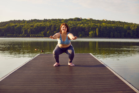 Happy plus size woman is training outdoors, by river or lake, performing squats. Overweight athlete is focused on exercise and smiling, embracing sport and fitness for weightloss.の写真素材
