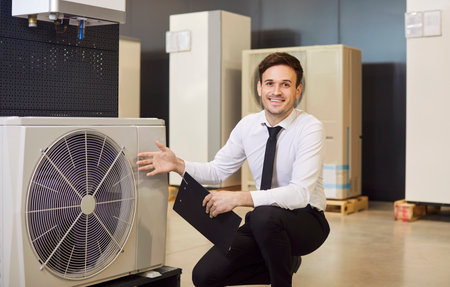 Happy, smiling man shop assistant with clipboard offers modern efficient sustainable ecologically friendly heat pump and HVAC split system for air conditioning available at their home appliances storeの写真素材