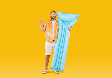 Funny young smiling man wearing beach clothes in sunglasses holding inflatable mattress isolated on studio yellow background. Happy tourist is going on summer holiday trip. Vacation concept.の写真素材