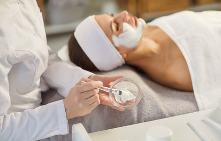 Side view of professional cosmetologist doing skincare treatment to young woman lying on couch. Beautician applying creamy facial mask with brush, performing rejuvenating procedure in beauty salon.の写真素材