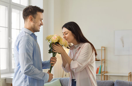 Young man congratulating on holiday happy girlfriend with bouquet of rose flowers. Smiling couple in love standing in living room at home. Valentines day and relationships concept.の写真素材