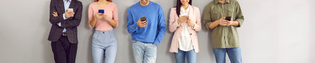 Happy young people using mobile phones while waiting by office wall. Several millennial men and women standing in row, holding smartphones, reading messages, or watching videos. Cropped shot, bannerの写真素材