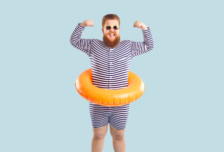 Bearded fat man with inflatable circle for swimming at waist shows off his biceps on pastel light blue background. Humorous redhead man in striped swimsuit in sunglasses shows his strengthの写真素材