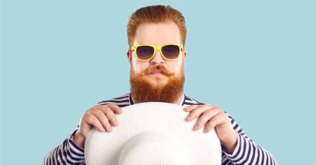 Portrait of funny chubby male summer vacationer pretending to be serious, isolated on light blue background. Close up of redhead bearded man in a striped sweater, sunglasses and with beach hat.の写真素材