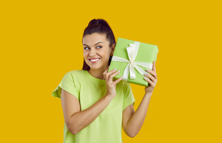 Happy woman shaking her present and trying to guess what's inside. Cheerful young girl standing isolated on yellow background, holding green gift box, listening to sound coming from inside and smilingの写真素材