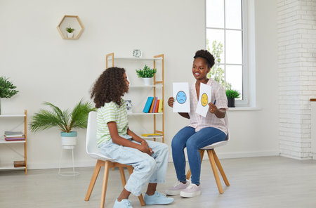 African american teenager girl is being consulted by a woman school psychologist in classroom. Teacher shows two emoticons cards sad and cheerful. Development of emotional intelligence teens concept.の写真素材