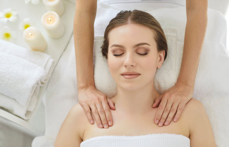 View from above of young woman getting professional masseuse is providing shoulder massage at luxurious spa, lying on couch, wrapped in white towel, feeling relaxed and comfortableの写真素材