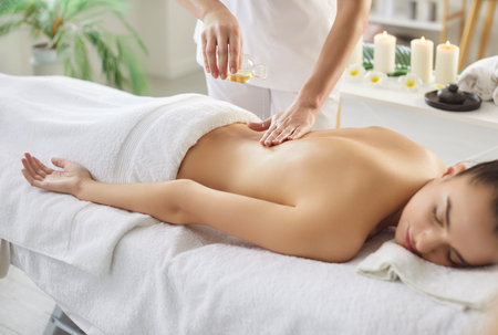 Therapist applying massage oil on womans back who is lying on massage table at spa, preparing for massaging with aromatherapy, covered with white towel. Side view. Body care conceptの写真素材