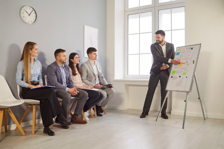 Confident businessman in suit standing near flip chart, presenting strategy to attentive colleagues sitting on chairs in office. Corporate coach explaining business plan during professional seminar.の写真素材