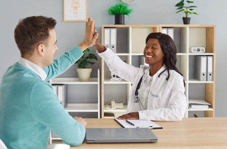 Friendly female doctor rejoicing at success in treatment giving high five to smiling man patient sitting at desk in medical clinic after medical examination. Health care and medicine concept.の写真素材