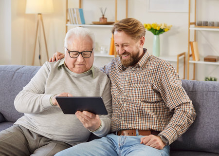 Elderly father and son enjoy online leisure at home on sofa, using tablet pc for internet entertainment, relaxing as family in a cozy setting with focus on connection and enjoyment.の写真素材