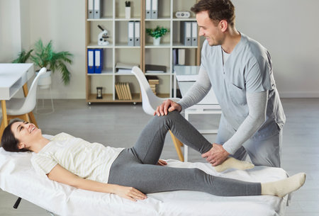 Man therapist or physiotherapist doing treatment of womans knee, providing medical care after physical injury. Pleased woman having rehabilitation session in private clinic, satisfied with resultsの写真素材