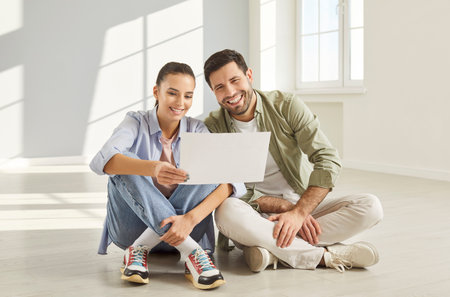 Happy young couple moving first new flat holding architectural plan document, resting seated on floor after relocation day, planning future, remodeling works, discuss repair ideas, renovationの写真素材