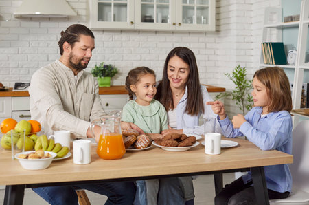 Portrait of smiling happy family of mother, father and two children having lunch sitting at table in kitchen. Young parents with cute kids eating cookies with orange juice at home.の写真素材