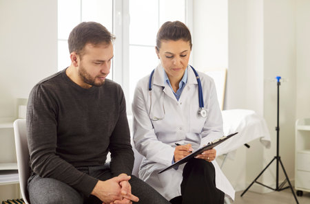 Portrait of a young man patient in medical office listening to a friendly female doctor holding report file with appointment and giving consultation during medical examination in clinic.の写真素材