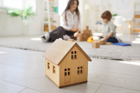 Closeup of little wooden toy house on floor in the living room with a family of young mother and little child boy playing together in background. Real estate and buying new home concept.の写真素材