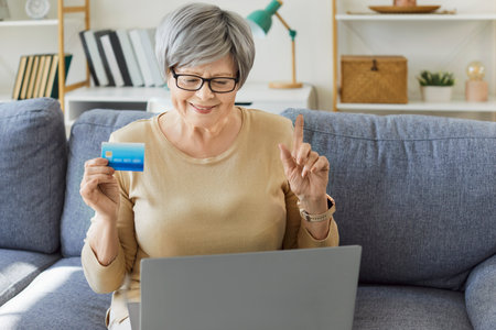 Happy elderly woman enjoys online shopping during leisure, making a purchase with a card on laptop at home, focusing on payment with excitement and engagement in the digital experience.の写真素材
