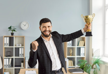 Portrait of excited happy smiling young man winner wearing formal clothes looking at camera holding the trophy award for success in business or for company best employee reward standing in office.の写真素材