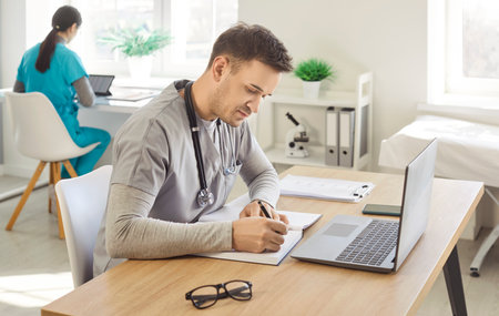 Young confident male doctor working at the desk with laptop on workplace doing paperwork in medical clinic or hospital with female nurse colleague. Medicine and health care concept.の写真素材
