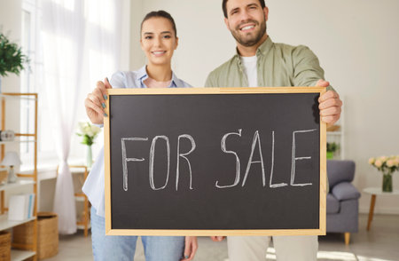 Smiling young couple standing in living room, holding chalkboard with handwritten message for sale. Happy man and woman presenting real estate offer, looking at camera, promoting home selling process.の写真素材