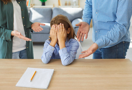 Upset child sitting at table with notebook, covering face with hands while his parents arguing or scolding in background. Father and mother criticizing kid because of educational or school problems.の写真素材