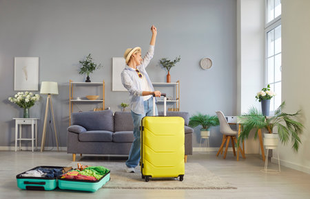 Positive woman dancing and celebrates while preparing for a summer vacation with a suitcase at home. The excitement of travel, trip planning, and holiday preparation fills the cozy domestic setting.の写真素材