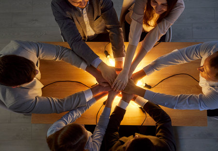Group of business people sitting in a circle, placing hands together in teamwork. Light bulb in background symbolizes innovation. Celebrating success in a corporate office environment.の写真素材