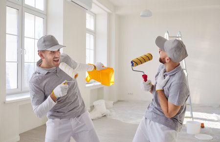 Funny skilled male painters having fun after finishing painting walls in apartment, fighting with paint rollers and shouting, doing house renovation, having fun with repair toolsの写真素材