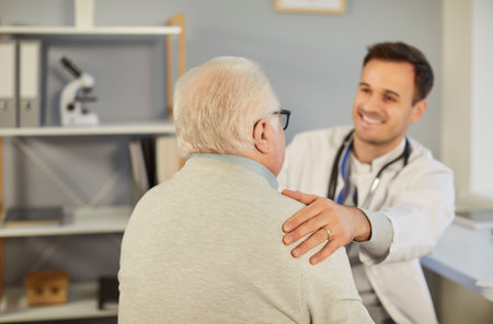 Senior sick man sitting in office during exam with young cheerful male physician. Doctor supporting his elderly patient sitting in medical clinic. Medicine and health care concept.の写真素材
