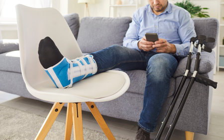 Disabled young man sitting at home on sofa in rehabilitation with crutches using mobile phone. Portrait of male person with injured leg after accident in plaster or splint with walking stick.の写真素材