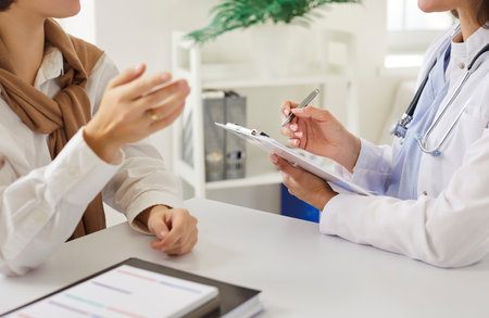Cropped view of female doctor attentively listening to woman patient and taking notes during consultation. Physician or cardiologist focusing on understanding and addressing patient health concerns.の写真素材