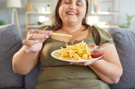 Plate of crispy french fries is in hands of cheerful woman comfortably seated on sofa at home. Face of woman radiates happiness as she indulges in junk food, demonstrating moment of pleasure.の写真素材