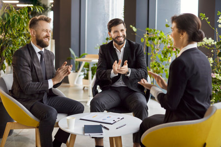 Business meeting in modern office. Two men and woman in suits engaging in productive discussion, smiling and clapping, celebrating successful deal, making agreement, expressing support and approval.の写真素材