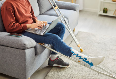 Broken leg or foot injury man, freelancer working with laptop online, crutches with bandaged leg at home, patient on rehabilitation period, painful swollen foot, severe fracture, consulting onlineの写真素材