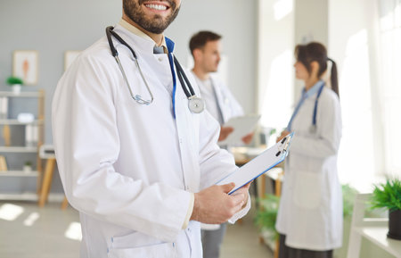 Cropped portrait of happy smiling young confident male doctor in white medical uniform standing in office. Man physician holding report file with appointment in clinic with colleagues in backgroundの写真素材