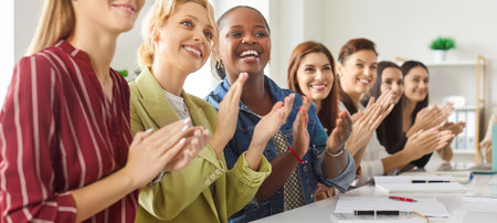 Cropped shot of smiling professional women sitting at long table and applauding to speaker or colleague. Happy female team engaged in conference, workshop or corporate training in office space. Bannerの写真素材