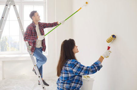 Young couple painting the wall of their new home holding paint rollers and standing on the ladder. Married man and woman doing repair renovation preparing to move into a new apartment.の写真素材