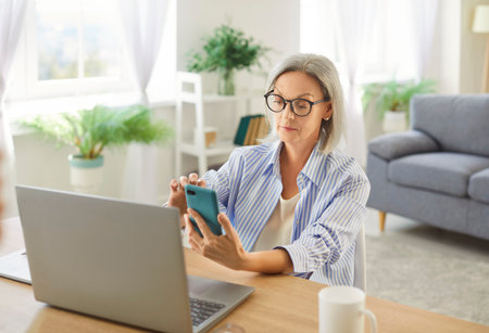 Senior mature woman working using laptop at home, sitting at table, aged female wearing glasses listening to lecture, watching webinar, studying online, learning language, phone using to screenの写真素材