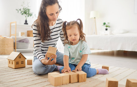Happy mother and cheerful cute preschool kid girl playing on carpet at home, stacking wooden cubes, constructing toy building blocks, smiling, enjoying leisure, playful babysitter entertainment gameの写真素材