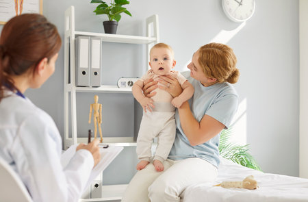 Young smiling mother sitting on couch in medical office, holding toddler baby in her hands during medical checkup with family doctor. Female pediatrician consulting caring mom raising her newborn kid.の写真素材