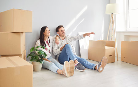 Portrait of a young happy couple man and woman sitting on floor in empty new apartment among cardboard boxes planning repair or design interior. Relocating, real estate, mortgage concept.の写真素材