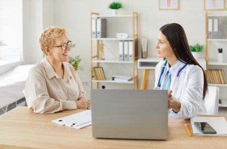Portrait of a young female doctor talking with sick mature smiling woman patient during consultation sitting at desk in clinic during medical examination. Health care and medicine concept.の写真素材